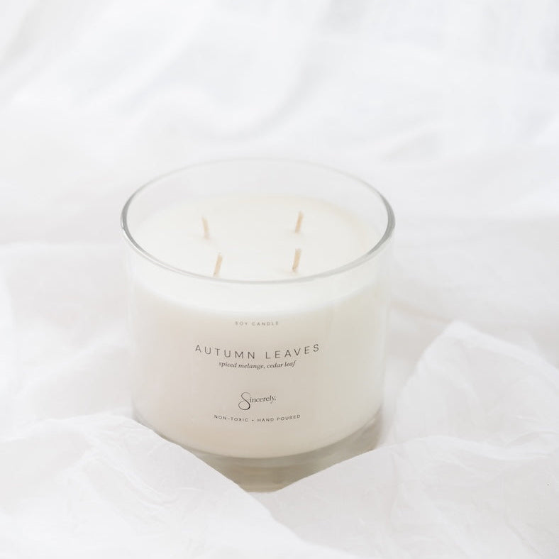 A 4-wick soy candle in a clear glass jar with white wax, labeled 'Autumn Leaves', styled on draped white fabric.