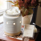 A white ceramic scented candle warmer with a lit interior sits on a brown book, alongside a small packaged wax melt labeled Harvest. A vase of burgundy flowers and a window are softly blurred in the background.