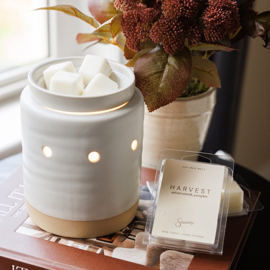 A white ceramic scented candle warmer with a lit interior sits on a brown book, alongside a small packaged wax melt labeled Harvest. A vase of burgundy flowers and a window are softly blurred in the background.