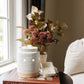 A white ceramic scented candle jar with a beige base sits on a dark round side table next to a window, styled with a book, a vase of dried flowers.