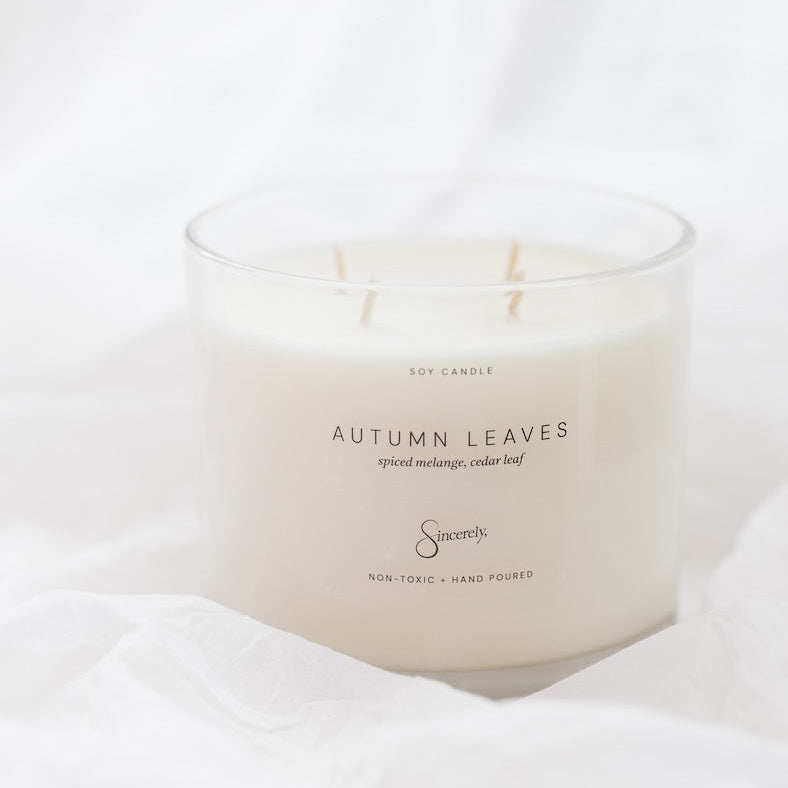 A white soy candle in a clear glass jar with four wicks, labeled 'AUTUMN LEAVES', set against a soft white fabric background.