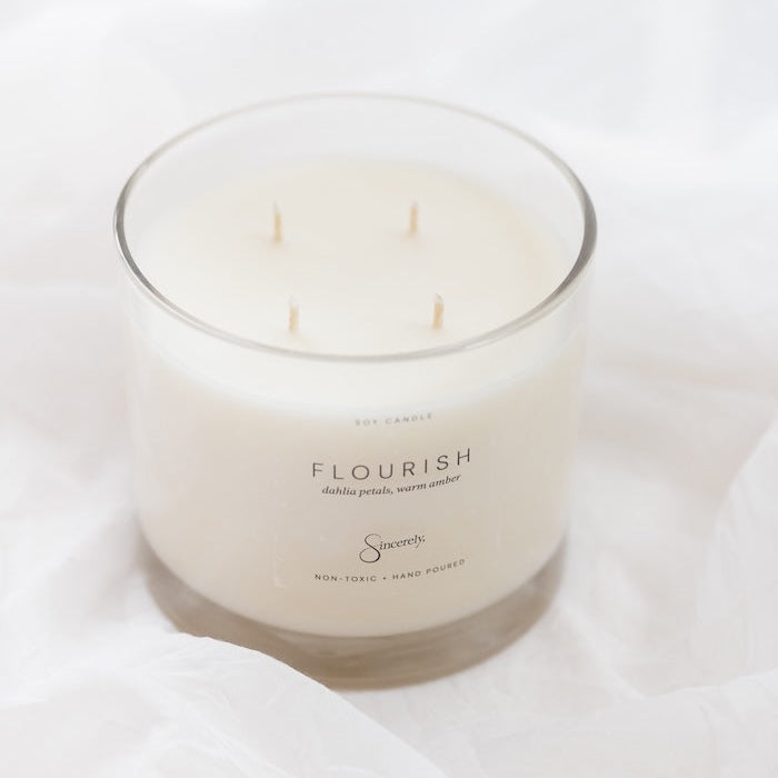 A 4-wick soy candle in a clear glass jar with cream-colored wax, resting on a soft, draped white fabric background. The label reads 'FLOURISH'