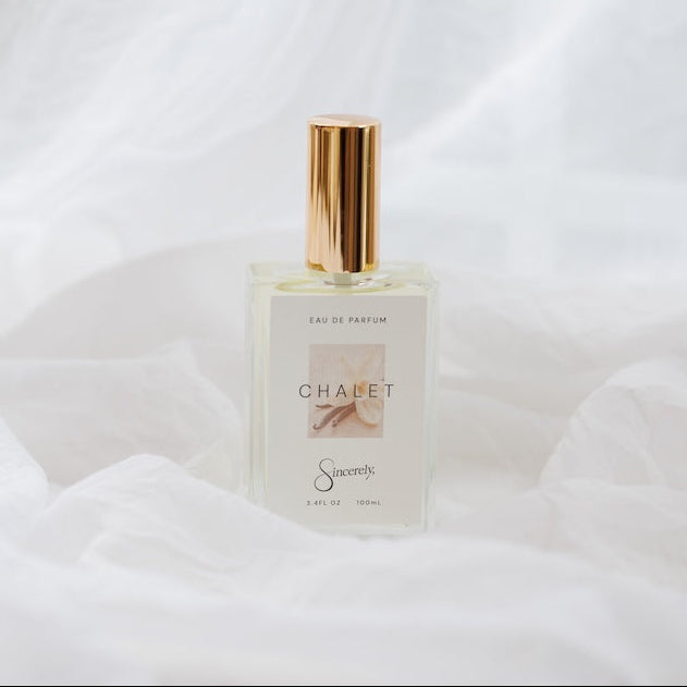 A clear glass perfume bottle with a gold cap and a minimalist white label reading 'CHALET' set against a draped white fabric background.