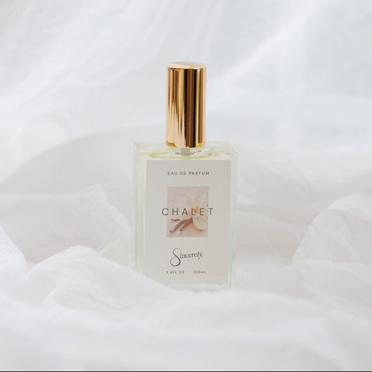 A clear glass perfume bottle with a gold cap and a minimalist white label reading 'CHALET' set against a draped white fabric background.