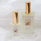 Two clear glass perfume bottles with gold caps and cream labels, one smaller in front of a larger bottle, set against a soft white draped fabric background. The labels shows the scent 'CHALET'.