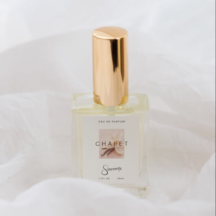 A small rectangular glass perfume bottle with a shiny metallic gold cap, labeled 'CHALET' set against a soft white draped fabric background.