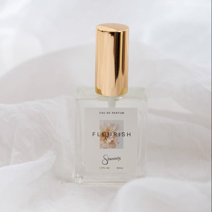 A clear glass perfume bottle with a shiny gold cylindrical cap, labeled 'FLOURISH' by the brand 'Sincerely', set against a draped white fabric background.