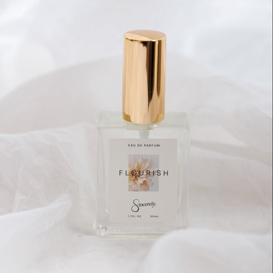 A clear glass perfume bottle with a shiny gold cylindrical cap, labeled 'FLOURISH' by the brand 'Sincerely', set against a draped white fabric background.