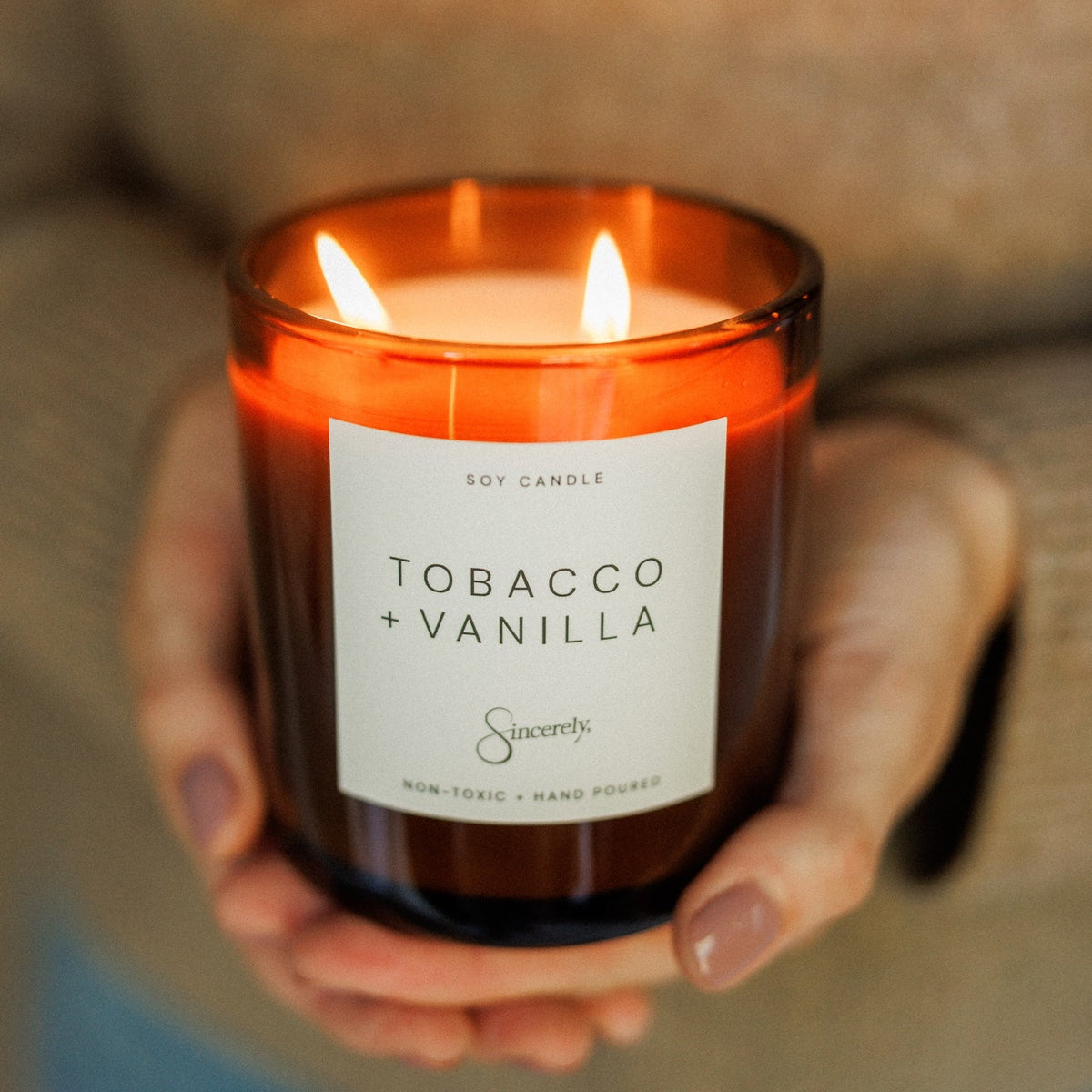 A person in a beige knit sweater holds a lit amber glass jar candle labeled 'TOBACCO + VANILLA' by Sincerely. The candle has two flames and is photographed in a cozy indoor setting with a blurred background.