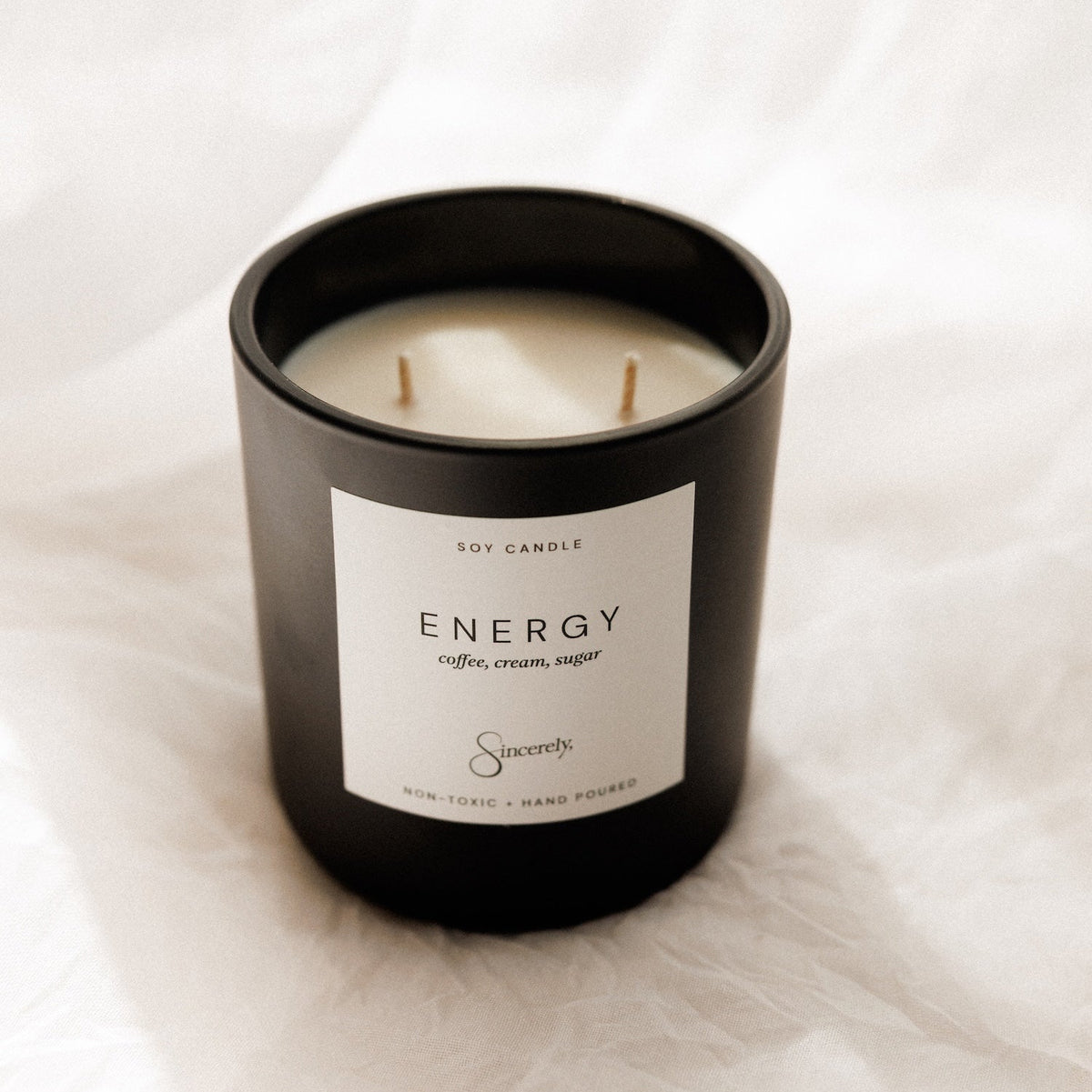 A black glass jar candle with two wicks and a white label reading 'ENERGY' placed on a softly draped white fabric background.