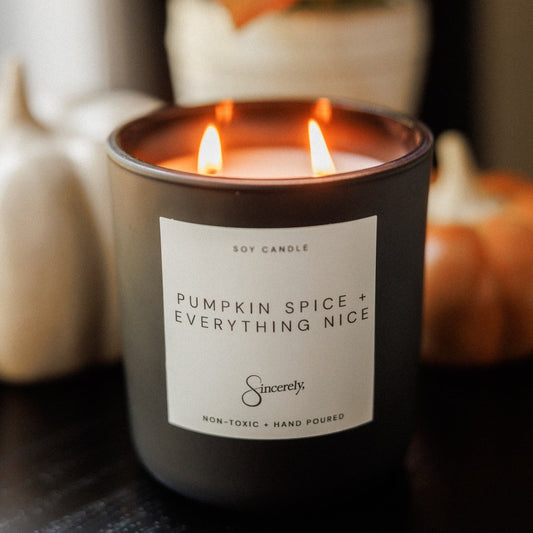 A lit black soy candle jar labeled 'Pumpkin Spice + Everything Nice' by Sincerely, set on a dark tabletop with blurred pumpkins and autumn leaves in the background.