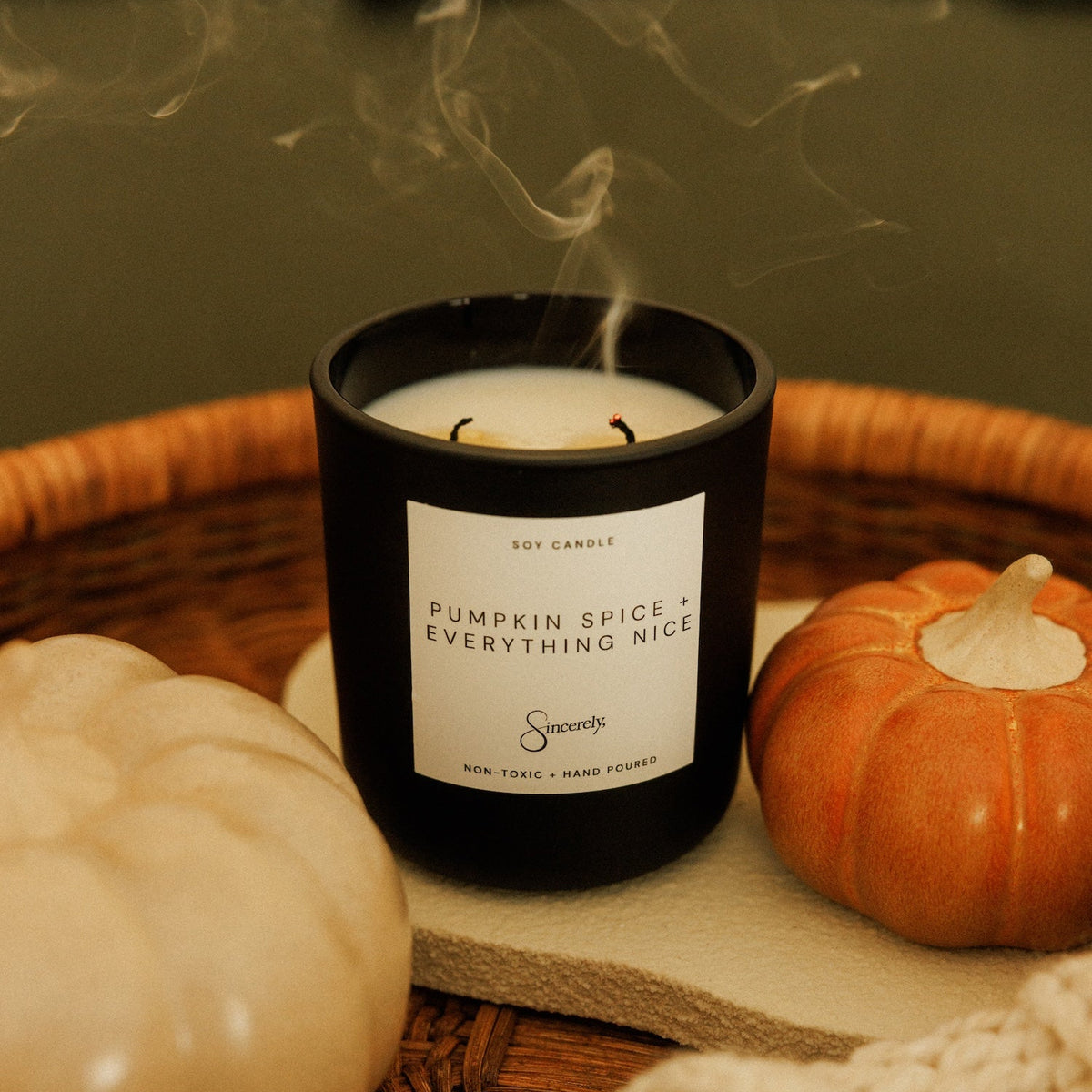 A black glass jar candle labeled 'Pumpkin Spice & Everything Nice' by Sincerely Candle, styled on a wicker tray with a small orange pumpkin, a cream-colored pumpkin, and a beige knit blanket.