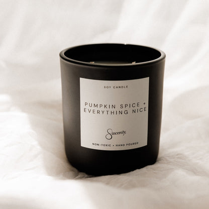 A black soy candle jar with a white label reading 'PUMPKIN SPICE + EVERYTHING NICE' and the brand name 'Sincerely' on a soft white fabric background.