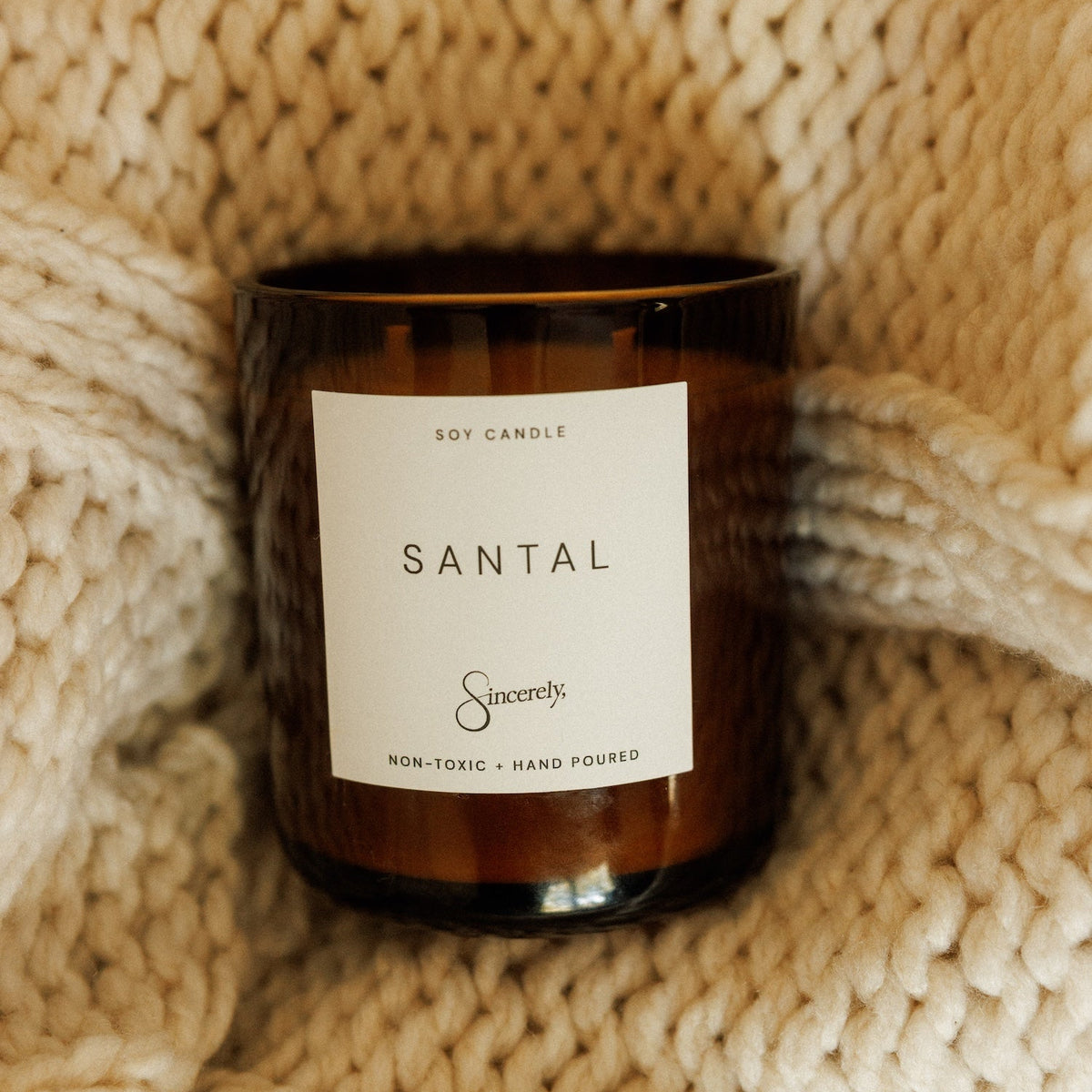 An amber glass soy candle with a white label reading 'SANTAL' and the brand 'Sincerely', styled on a chunky cream knit blanket.