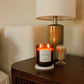A lit candle in a dark amber glass jar sits on a wooden bedside table next to a lamp and a small decorative glass container in a softly lit bedroom setting.