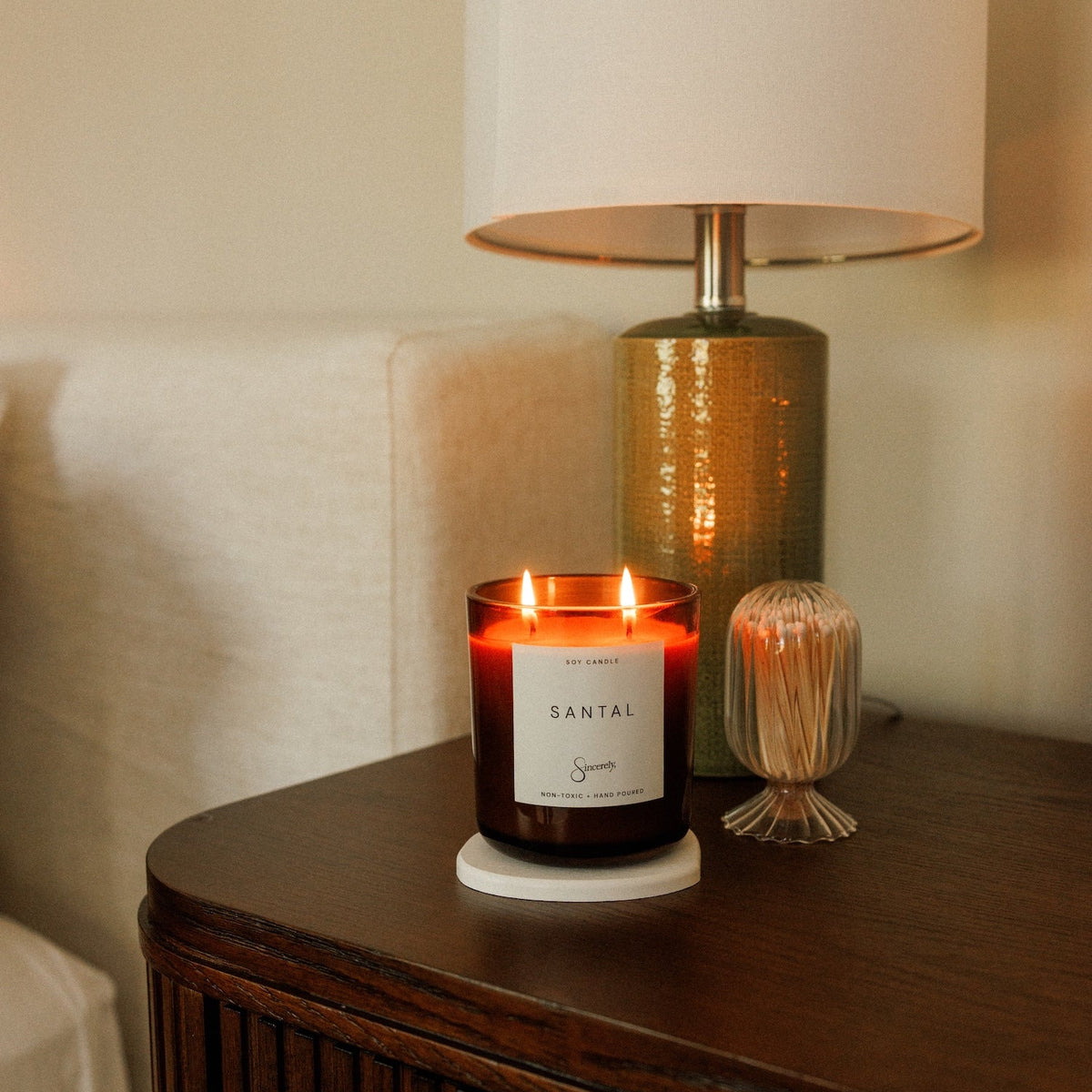 A lit candle in a dark amber glass jar sits on a wooden bedside table next to a lamp and a small decorative glass container in a softly lit bedroom setting.
