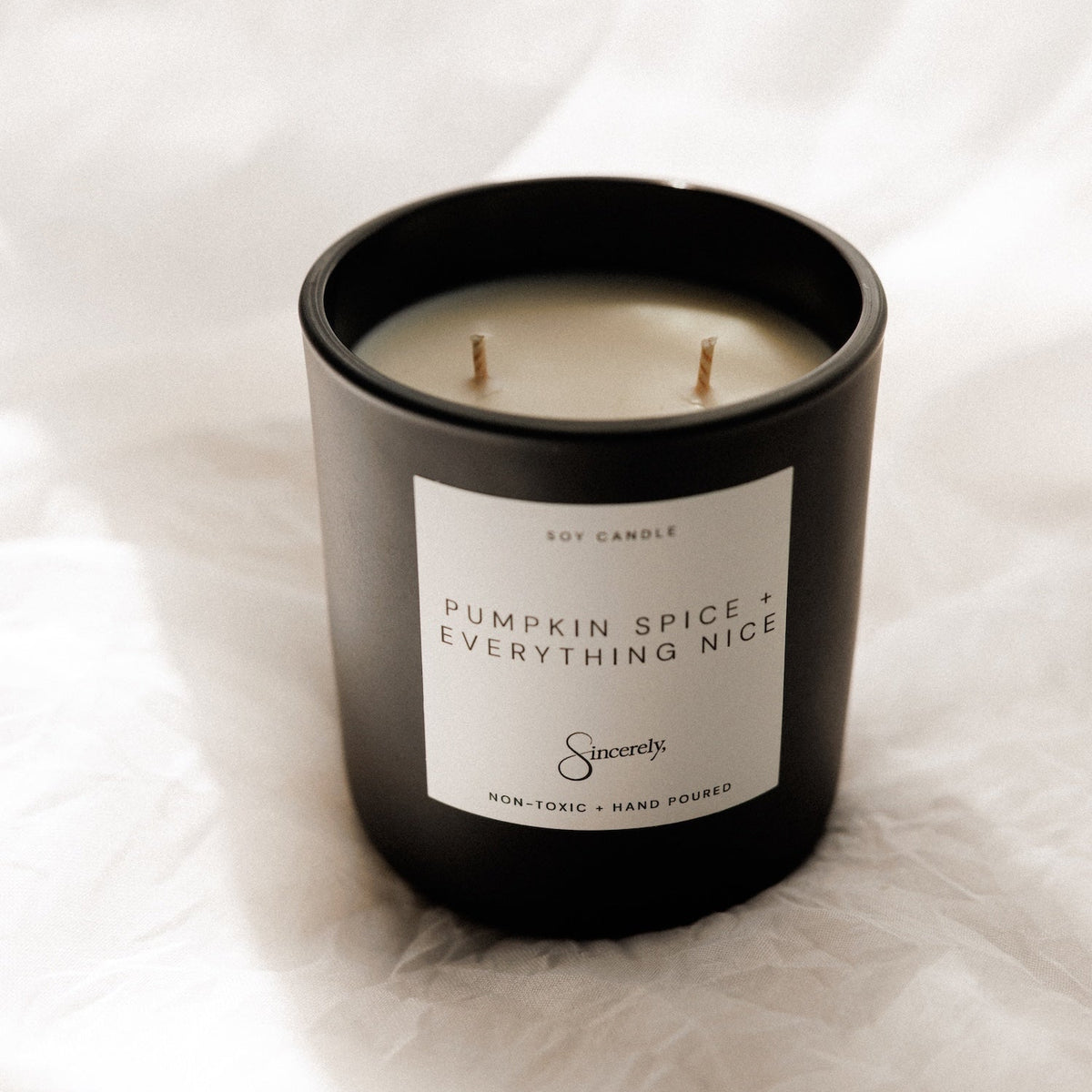 A black two-wick soy candle with a white label reading 'Pumpkin Spice + Everything Nice' and the Sincerely Candle logo, set against a draped white fabric background.