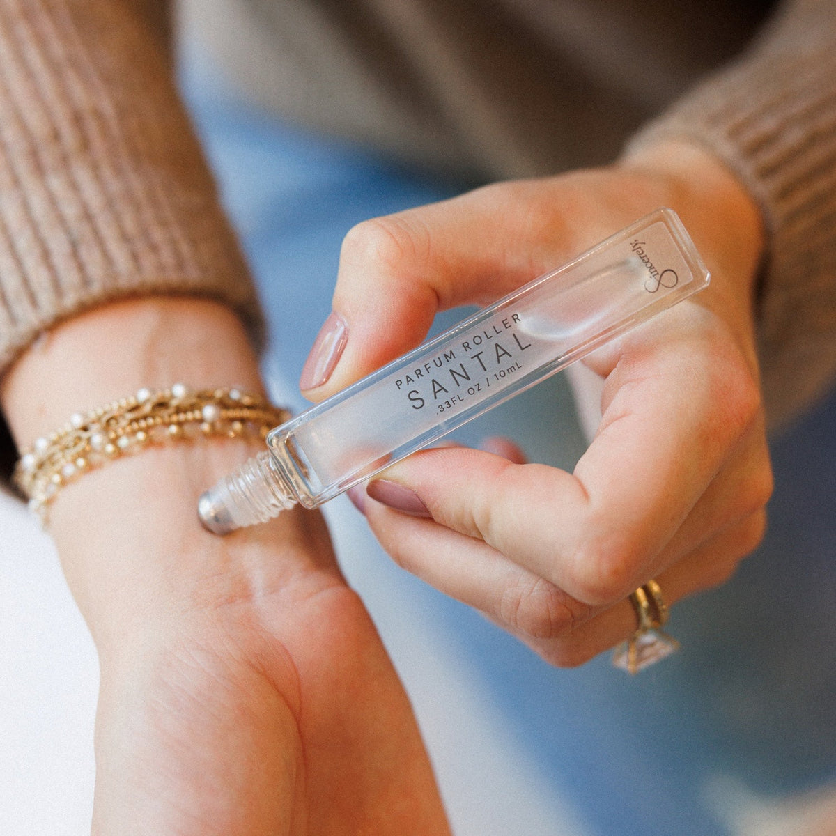 A close-up of a person's hand holding a clear glass rollerball bottle labeled 'SANTAL' while applying perfume to their wrist 
