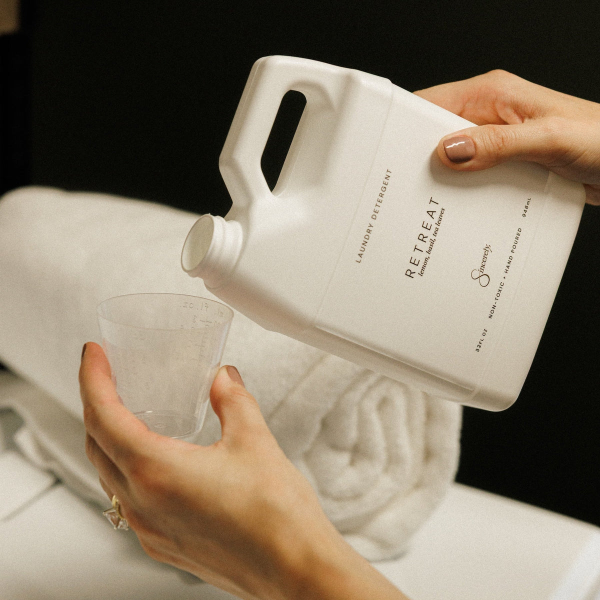A person pouring a white liquid from a large white plastic jug labeled 'RETREAT' into a clear measuring cup, with rolled white towels in the background.