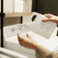 A person pouring liquid from a white plastic bottle labeled 'RETREAT' into a small clear cup, set in a bright laundry room with a white washing machine in the background.