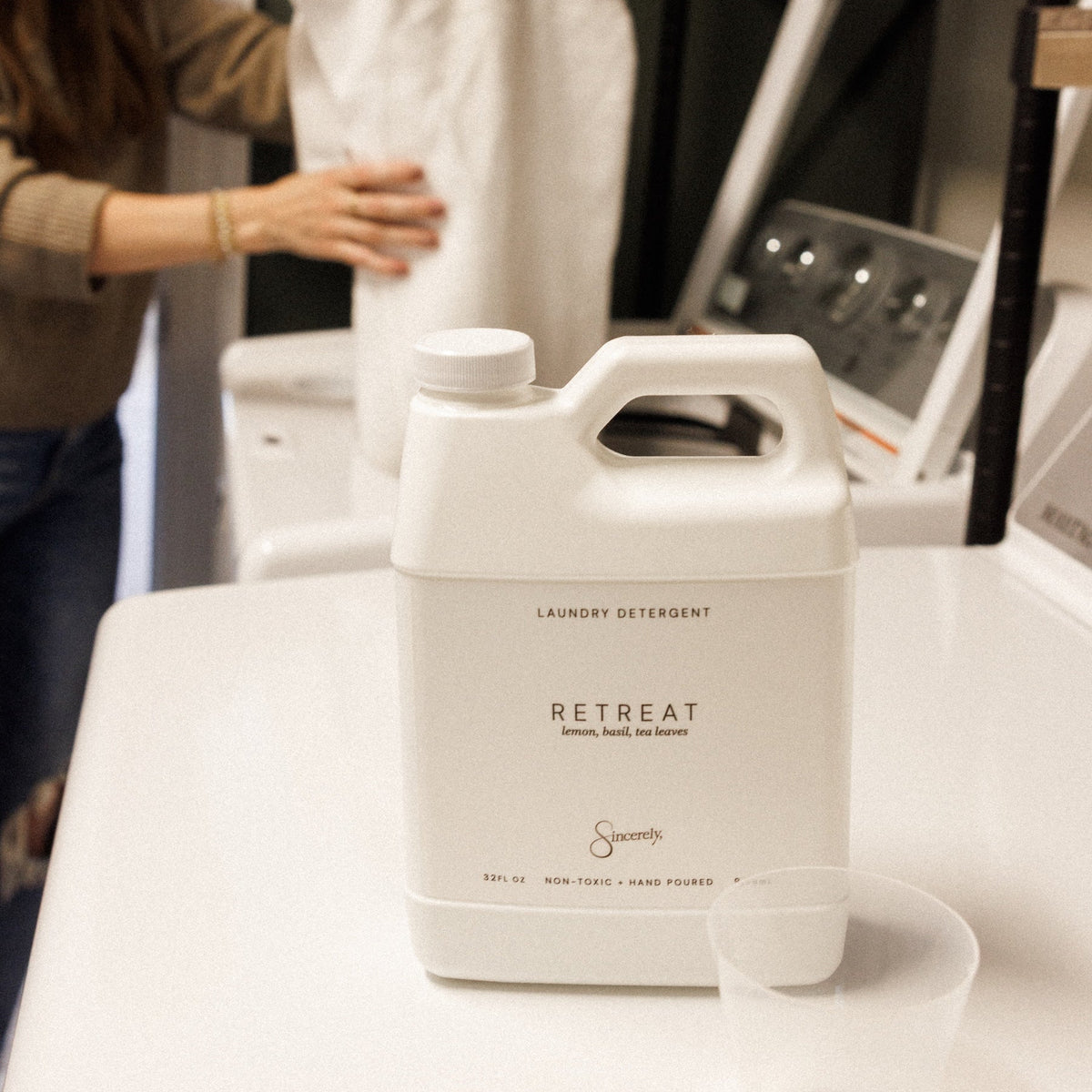 A white plastic bottle of Retreat laundry detergent sits on a white counter in a laundry room, with a person in the background handling a white garment and a small clear measuring cup in the foreground.