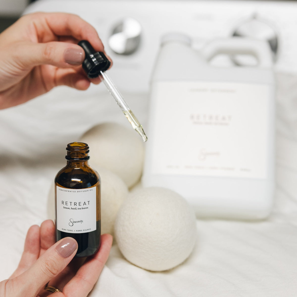 A person holds a small amber bottle with a dropper and a white jug labeled RETREAT in a spa-like setting on white linens.