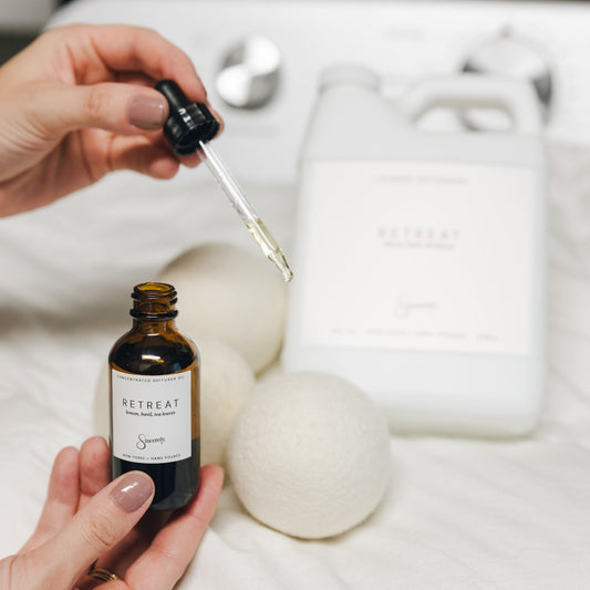 A person holds a small amber bottle with a dropper and a white jug labeled RETREAT in a spa-like setting on white linens.