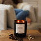 A black glass candle labeled 'ENERGY' with two lit wicks sits on a wooden table in a cozy living room setting, with coffee beans and matches in front and a blurred person drinking in the background.