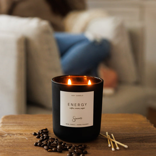 A black glass candle labeled 'ENERGY' with two lit wicks sits on a wooden table in a cozy living room setting, with coffee beans and matches in front and a blurred person drinking in the background.