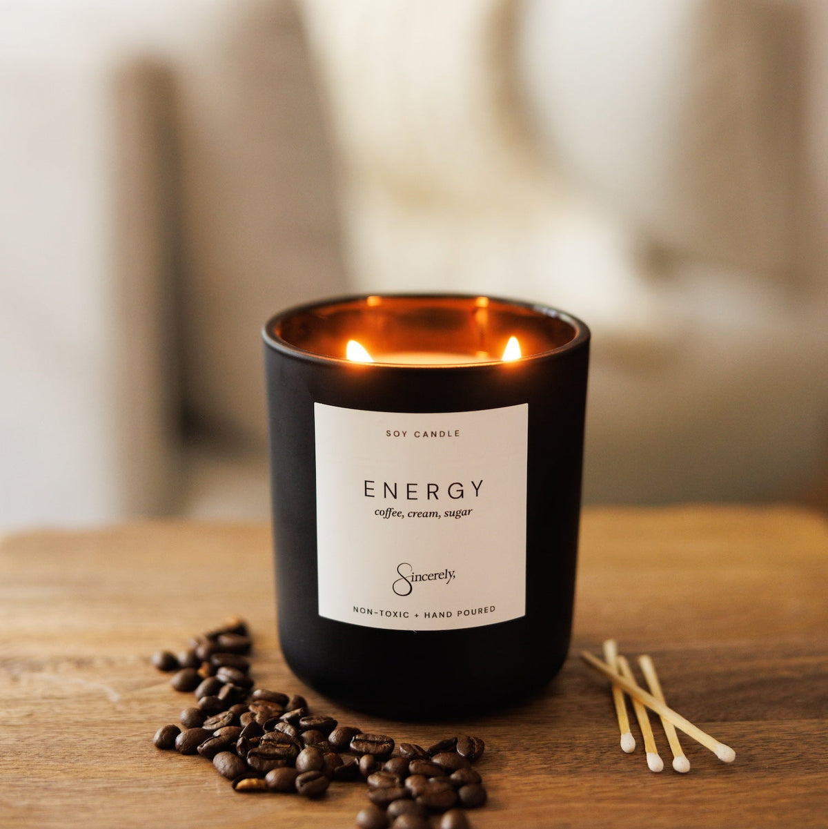 A black glass jar candle with two lit flames sits on a wooden table, surrounded by coffee beans and wooden matches. The candle features a white label with the scent name 'ENERGY' and the brand Sincerely