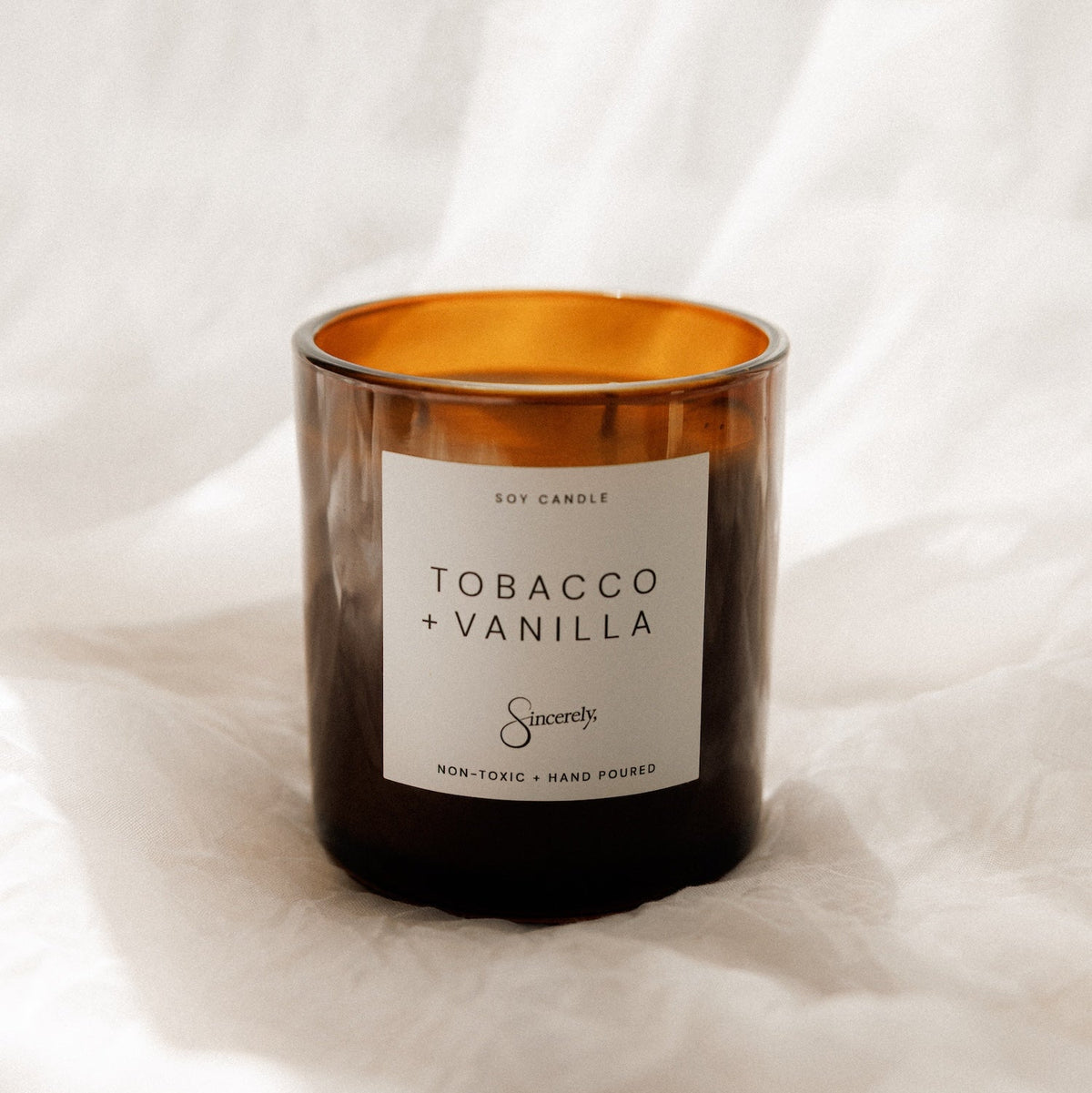 An amber glass jar candle with a white label reading 'TOBACCO + VANILLA' and the brand name 'Sincerely' on a soft white fabric background.