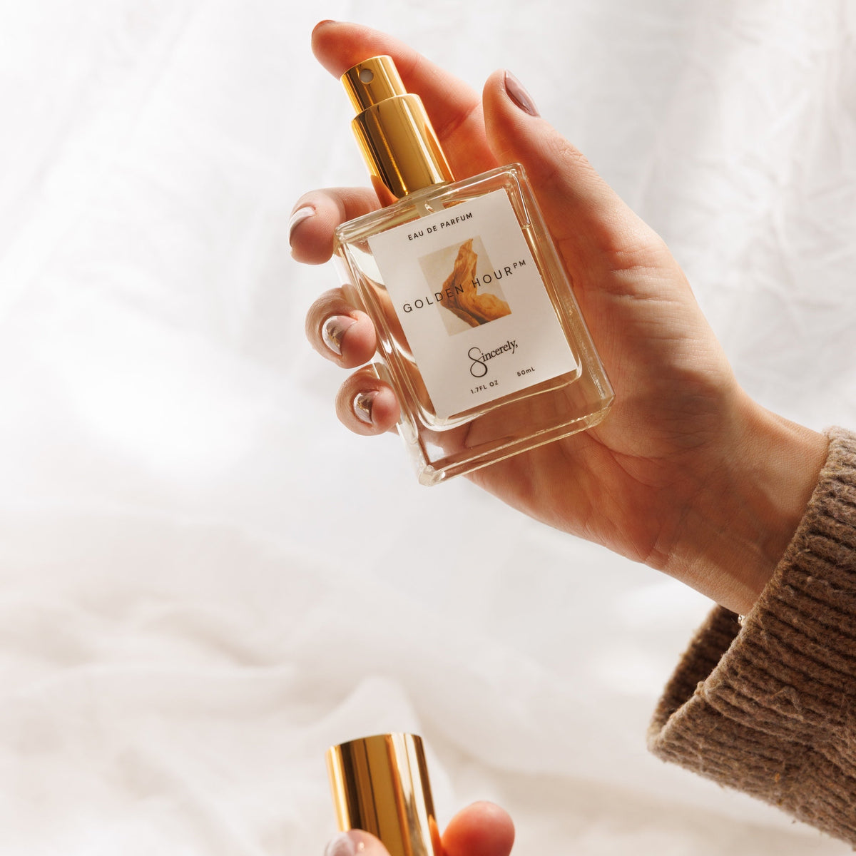 A person holds a clear glass perfume bottle with a gold spray top and a white label in one hand, while removing the gold cap from the other hand, set against a soft white draped fabric background.