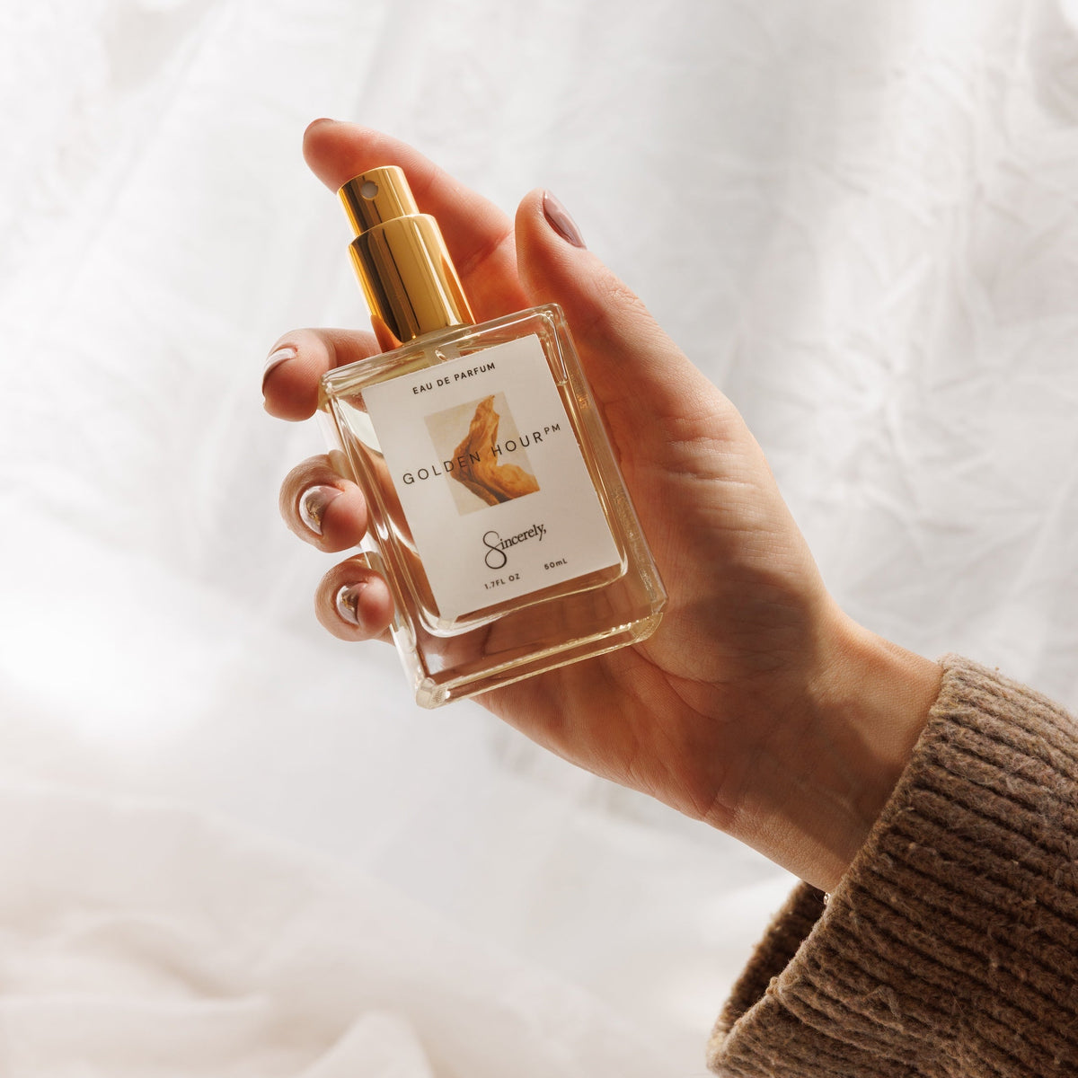 A hand holding a square glass perfume bottle with a gold spray top and a white label. The hand is wearing a brown ribbed knit sweater sleeve, set against a soft, draped white fabric background.