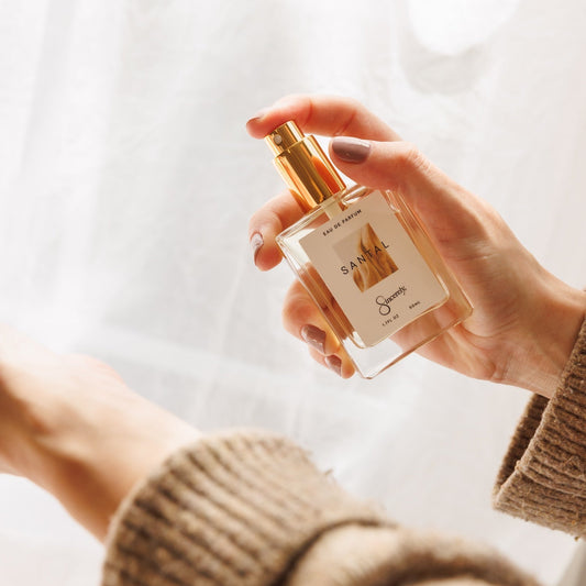 A hand holding a clear glass perfume bottle with a gold spray top and a cream label, photographed against a soft white curtain background. The person is wearing a beige ribbed sweater.