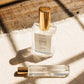 A clear glass perfume bottle with a gold cap and a matching small travel atomizer bottle, both labeled COVE, placed on a textured cream fabric with warm sunlight and window shadow patterns.