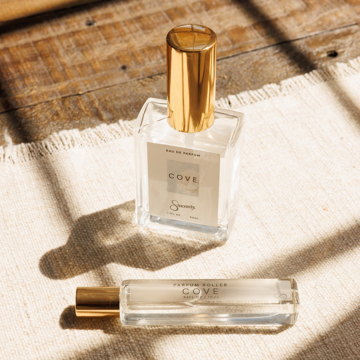 A clear glass perfume bottle with a gold cap and a matching small travel atomizer bottle, both labeled COVE, placed on a textured cream fabric with warm sunlight and window shadow patterns.
