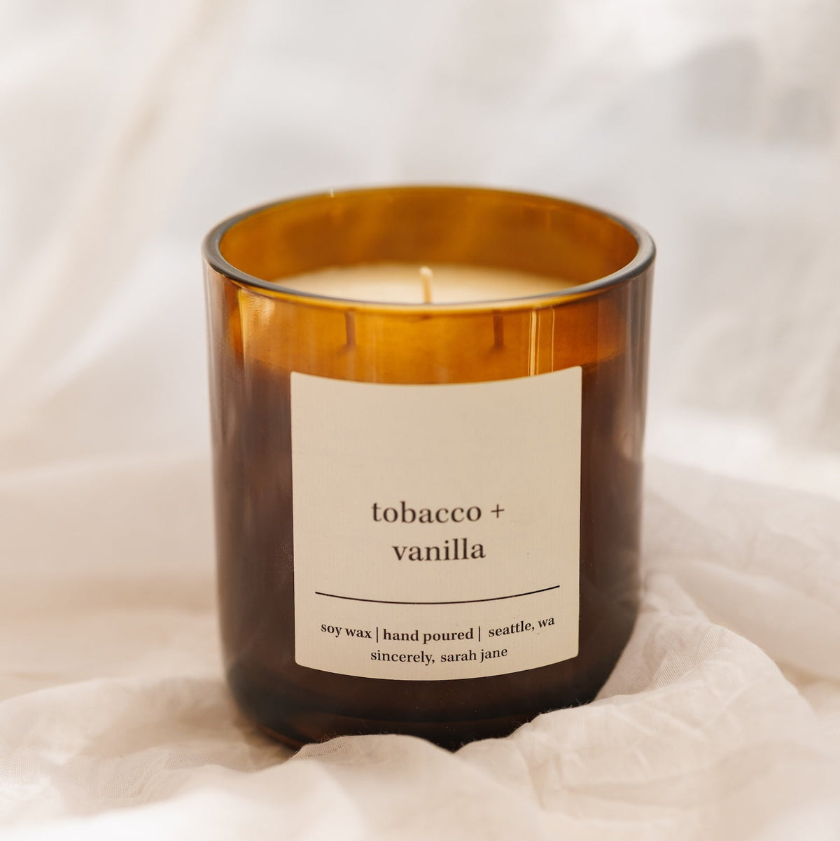 An amber glass jar 3-wick candle with a white label reading 'tobacco + vanilla' on a soft white fabric background.