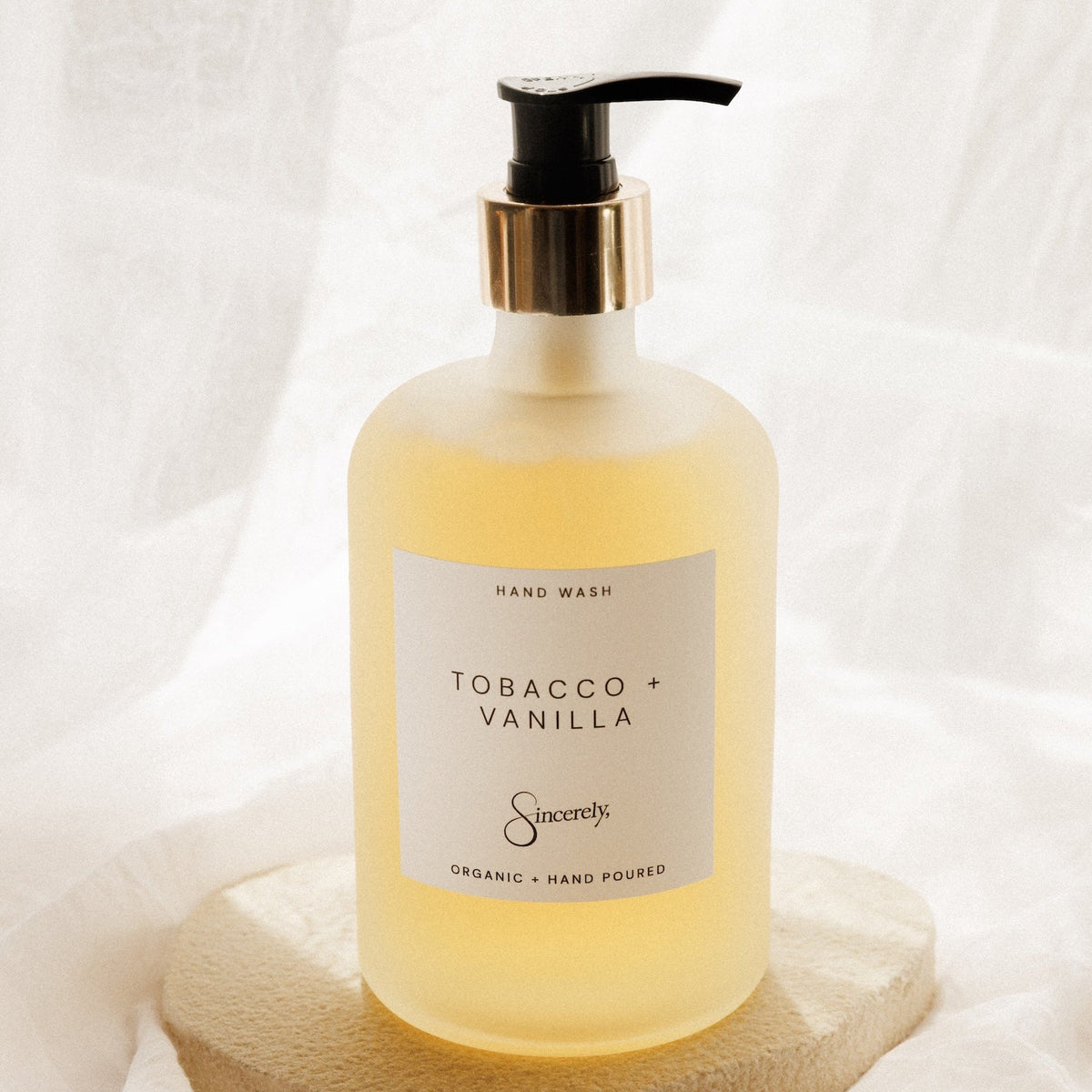A frosted glass hand wash bottle with a black pump and gold collar, filled with a pale yellow liquid. The bottle has a white label reading 'TOBACCO + VANILLA' and is displayed on a textured stone slab against a draped white fabric background.