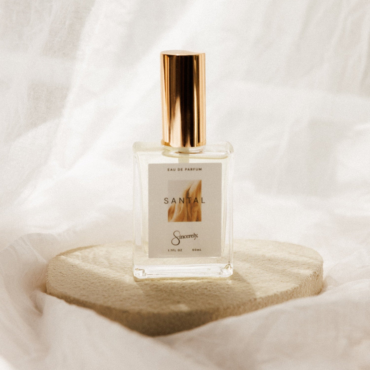 A clear rectangular perfume bottle with a metallic gold cap and a white label reading 'Santal', displayed on a round beige stone pedestal against a softly draped white fabric background.