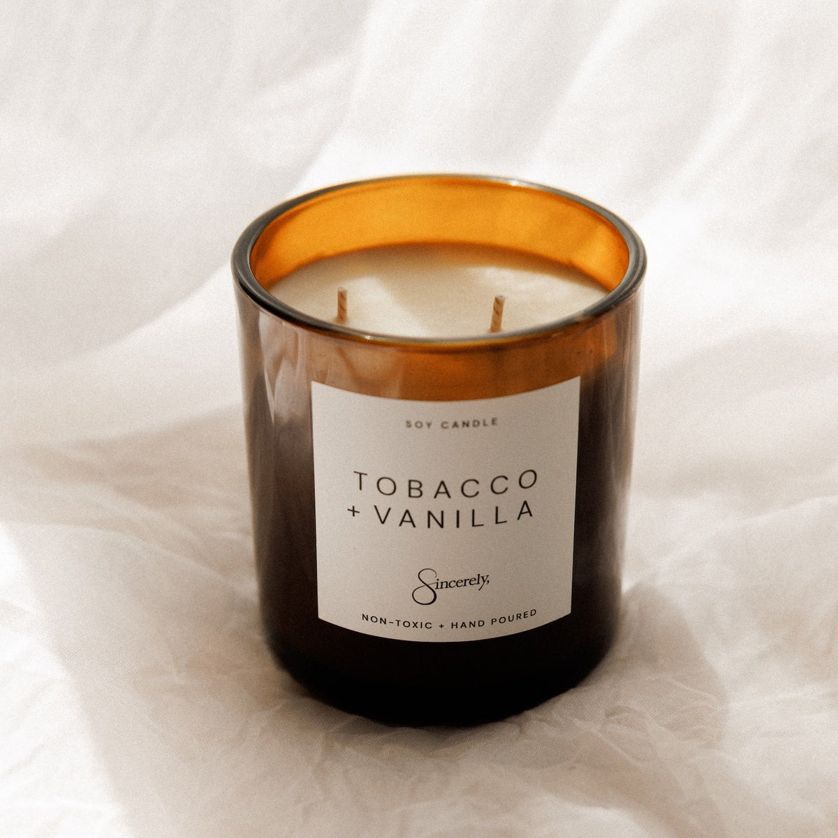 A two-wick soy candle in an amber glass jar with a white label reading 'TOBACCO + VANILLA' and the brand 'Sincerely', set against a draped white fabric background.