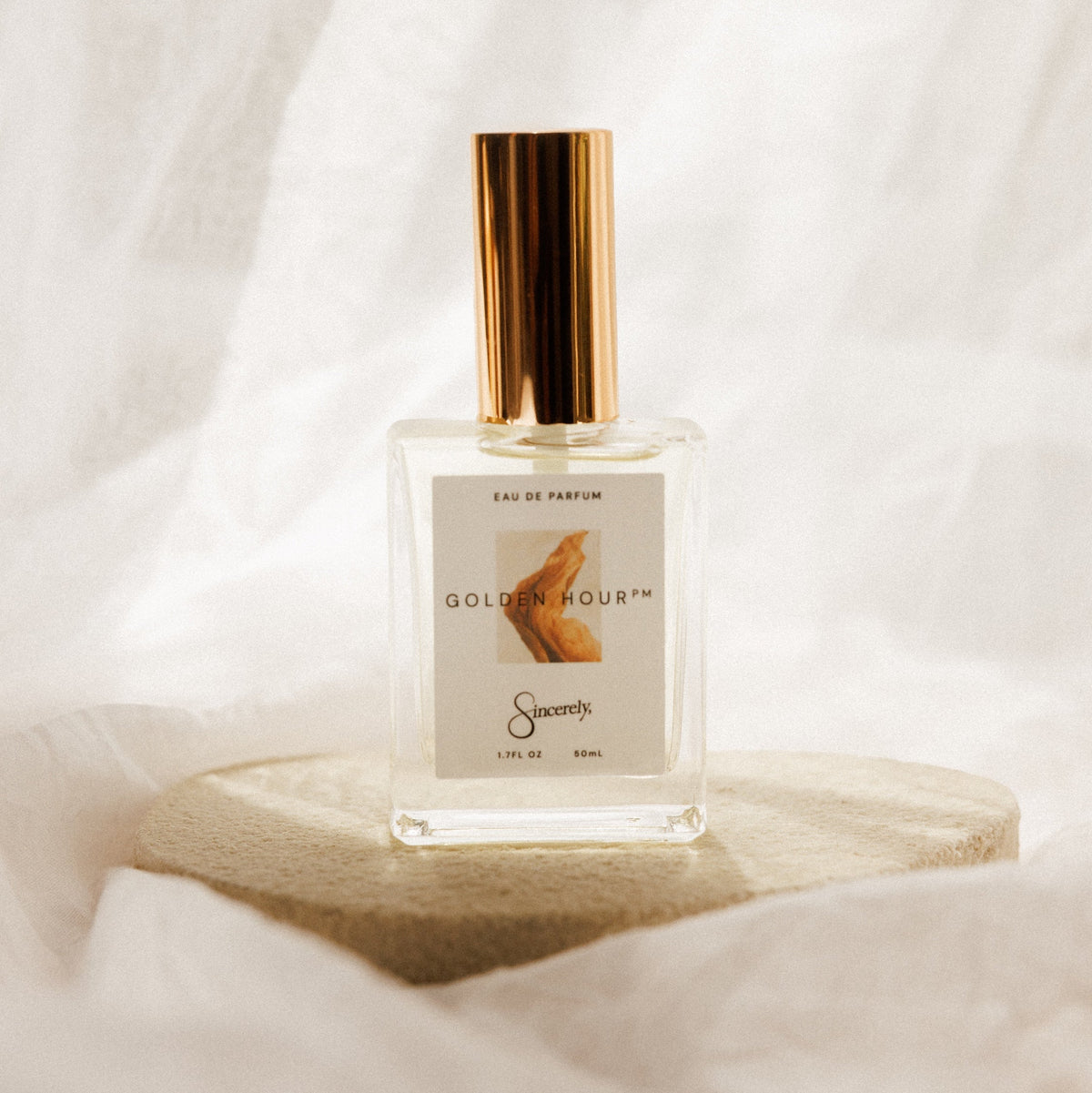 A clear glass perfume bottle with a shiny gold cap, labeled 'GOLDEN HOUR' by 'Sincerely', displayed on a round beige stone pedestal against a draped white fabric background.