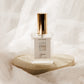 A clear glass perfume bottle with a metallic gold cap and a white label reading 'COVE', displayed on a round beige stone pedestal against a draped white fabric background.