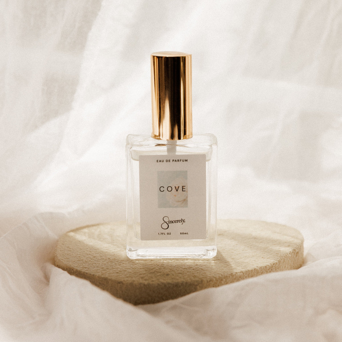 A clear glass perfume bottle with a metallic gold cap and a white label reading 'COVE', displayed on a round beige stone pedestal against a draped white fabric background.