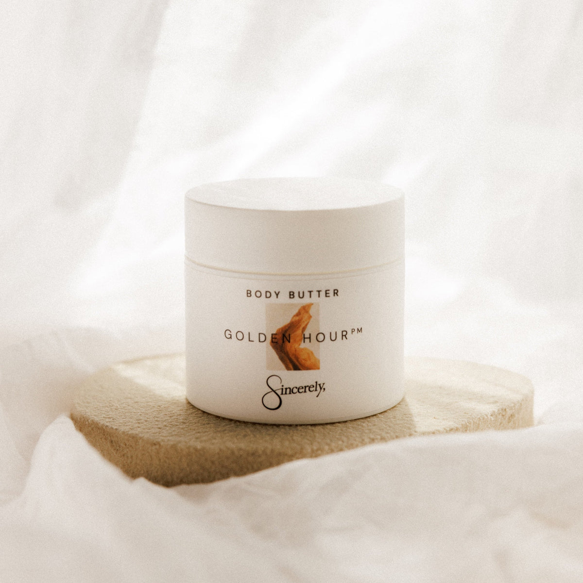 A white jar of Sacrety Golden Hour Body Butter sits on a round beige stone tray against a soft, draped white fabric background.