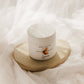 A white jar of Golden Hour PM Body Butter sits on a textured beige coaster, surrounded by soft, draped white fabric.