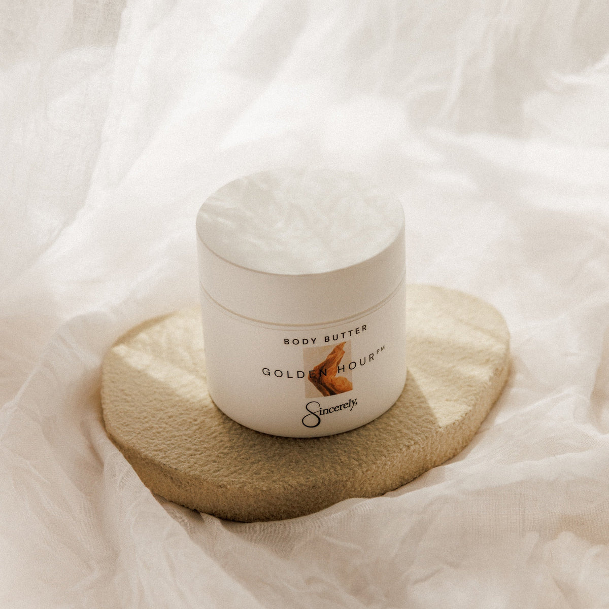 A white jar of Golden Hour PM Body Butter sits on a textured beige coaster, surrounded by soft, draped white fabric.