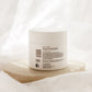 A white cosmetic jar with a white lid, showing the back label, resting on a textured beige stone slab against a soft, draped white fabric background.