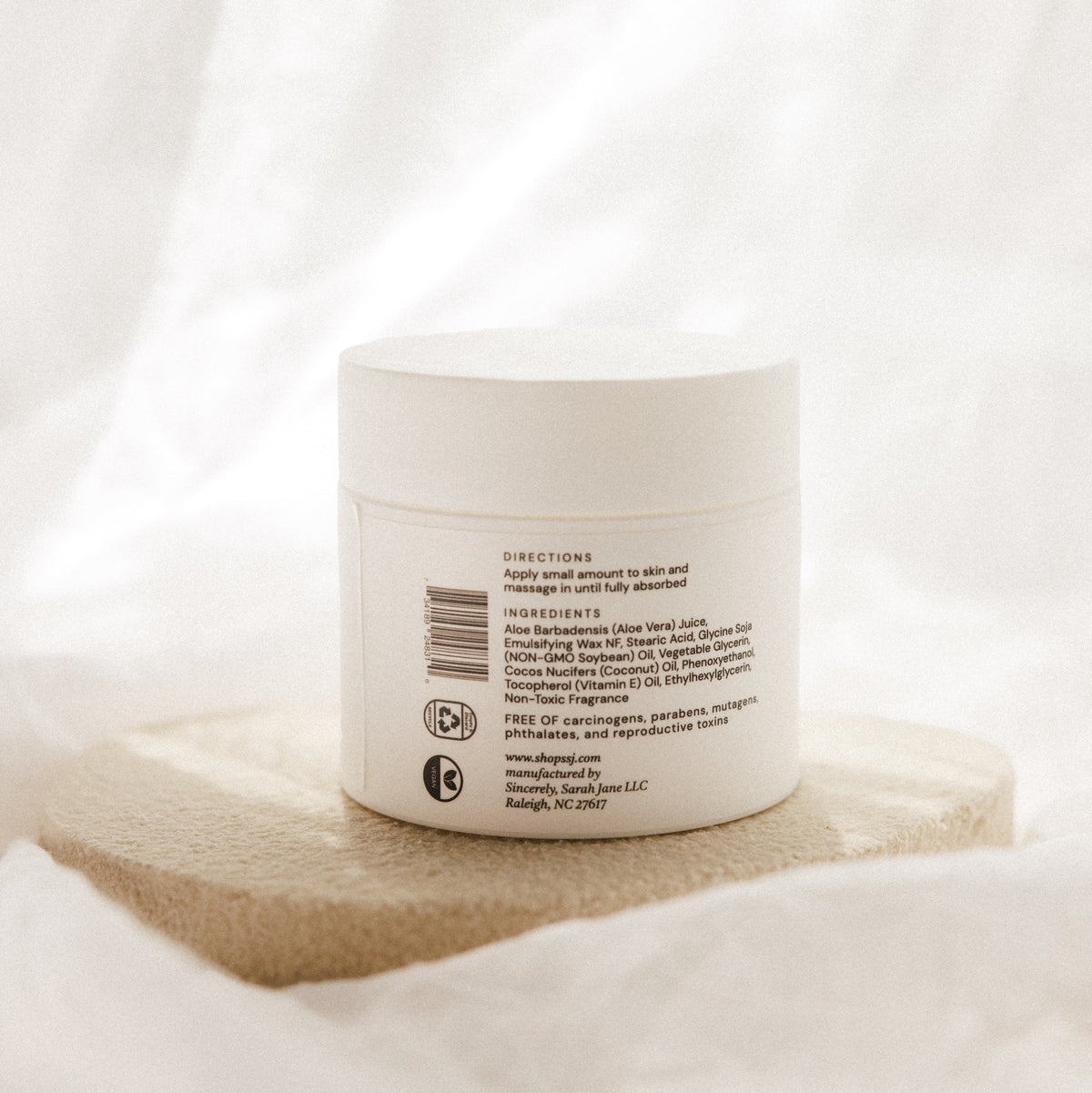 A white cosmetic jar with a white lid, showing the back label, resting on a textured beige stone slab against a soft, draped white fabric background.
