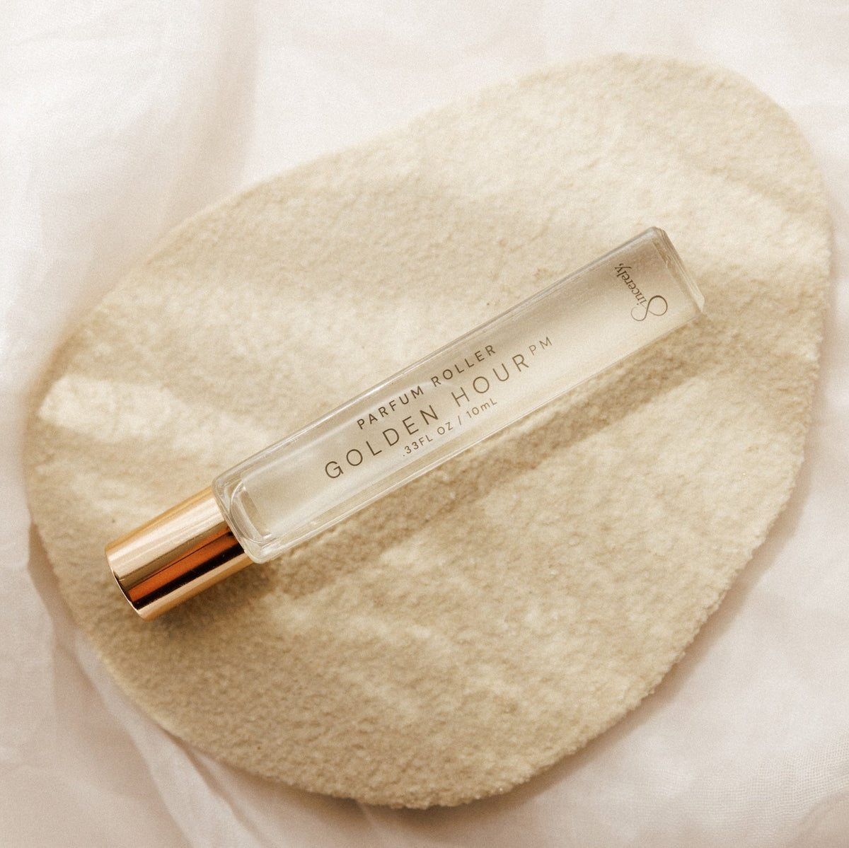 A clear glass perfume roller bottle with a metallic gold cap, labeled 'GOLDEN HOUR PM' resting on a textured beige fabric pad over a soft white background.