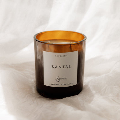 An amber glass soy candle with a white label reading 'SANTAL' and the brand 'Sincerely', set against a draped white fabric background.